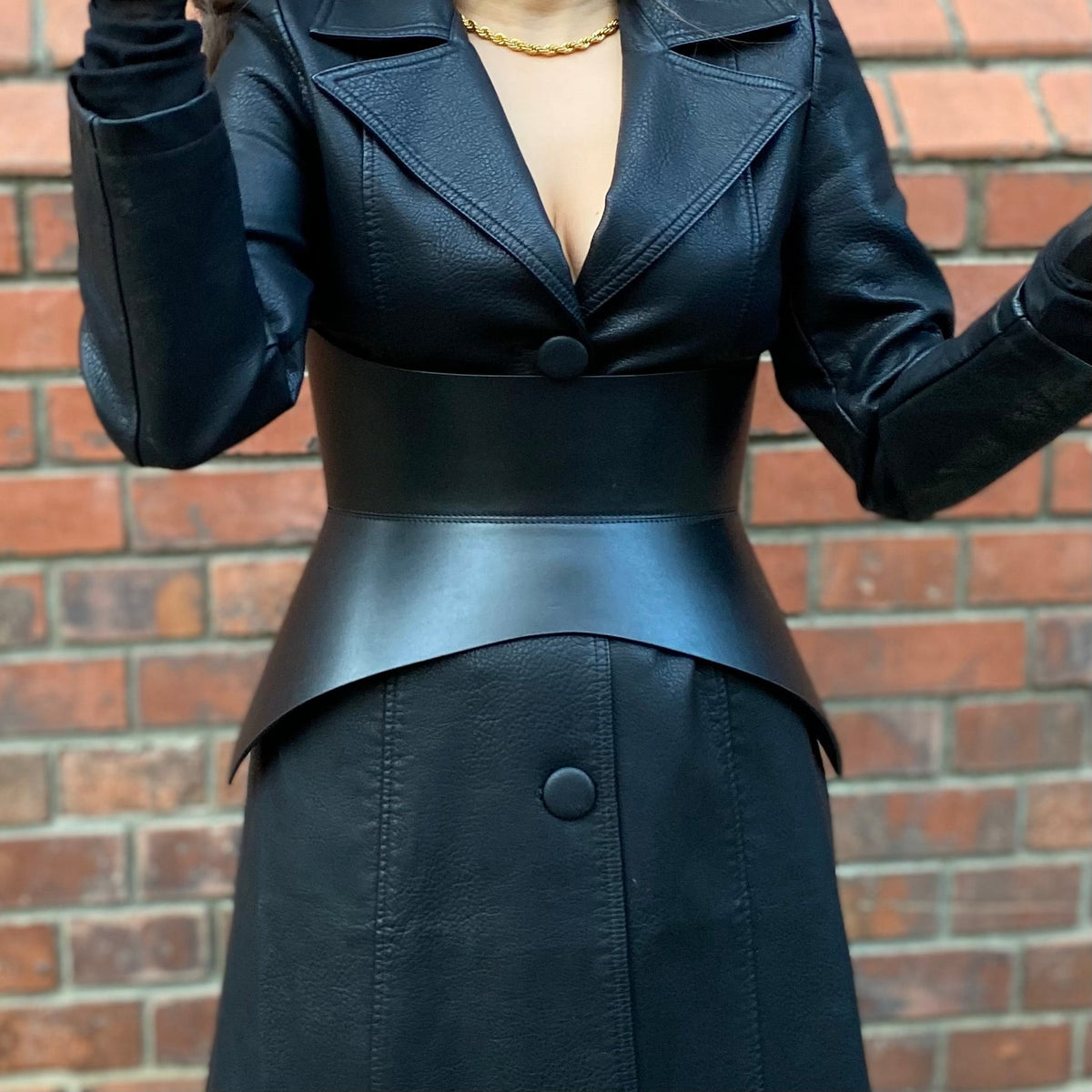 Aria Margo black leather corset belt worn over a matching black leather coat against a red brick wall background.