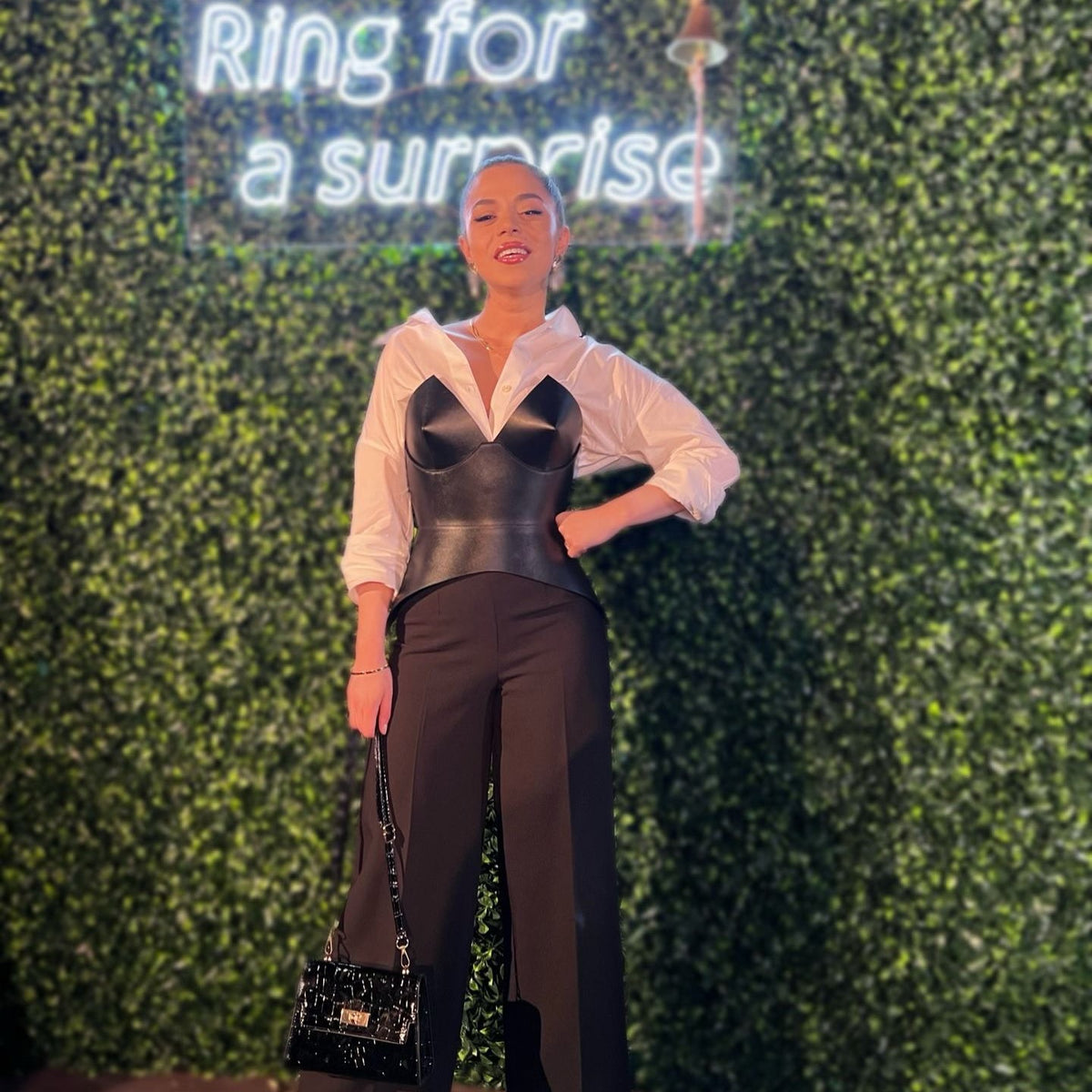 Aria Margo black leather full body corset belt worn over a white shirt and black trousers against a green wall.