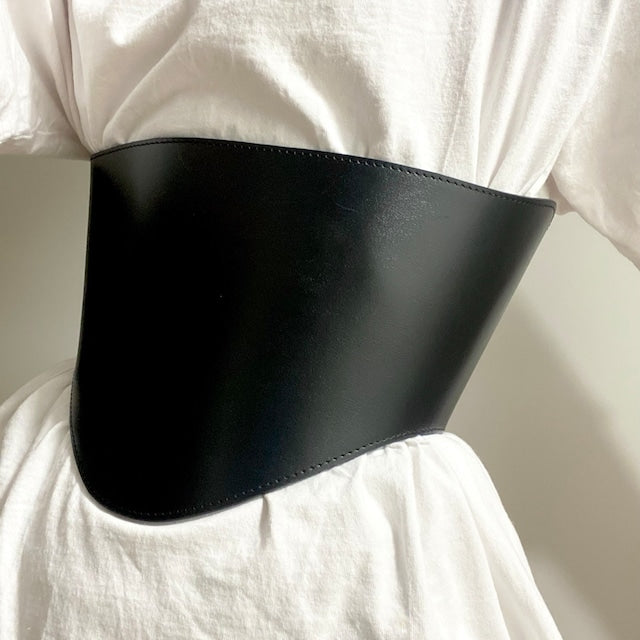 Aria Margo black leather under-bust belt with a curved design worn over a white top to define the waist.