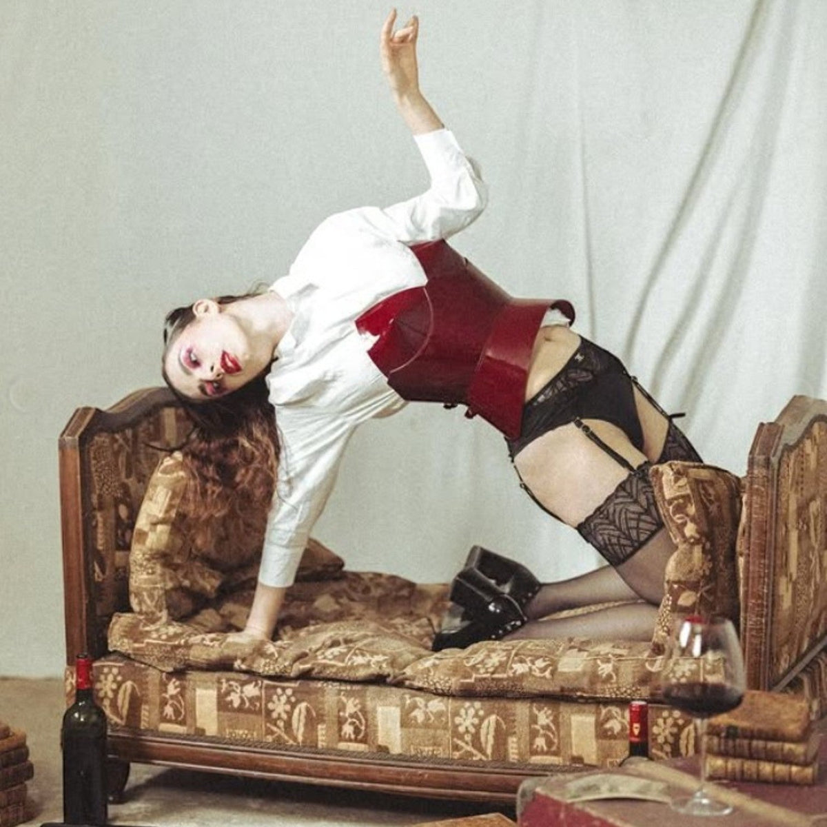Aria Margo burgundy patent leather round body corset worn over a white shirt by a model on a vintage sofa.