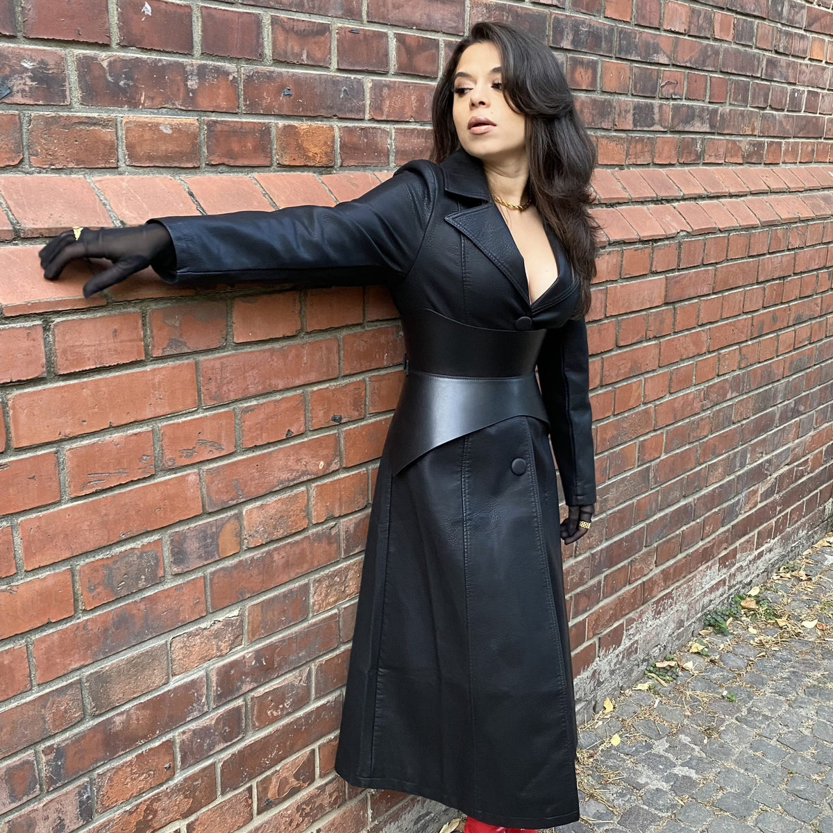 Aria Margo black leather Hallo corset belt worn over a matching black leather trench coat against a brick wall