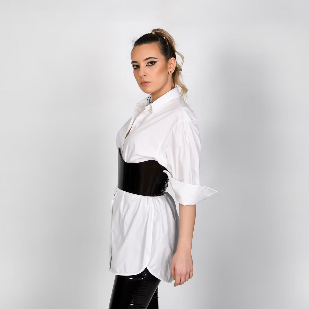 Aria Margo Crown under-bust belt in high-shine patent black leather worn over a crisp white button-down shirt.