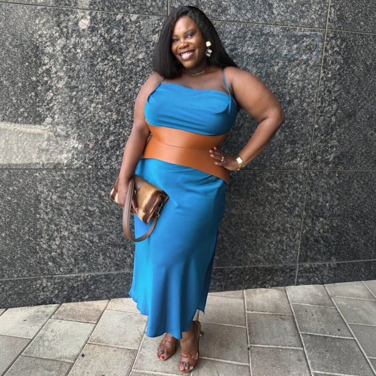 Aria Margo Test brown leather corset belt worn over a blue midi dress by a smiling woman against a stone wall.