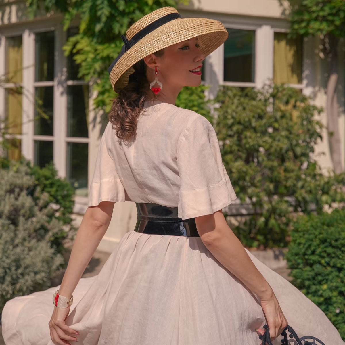 Aria Margo slim black patent leather corset belt worn over a light beige dress by a woman in a straw hat